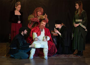 A production shot of the show: Henry VIII holds court sat on a throne
