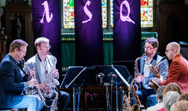 Four saxophonists performing, with music stands and grand purple banners